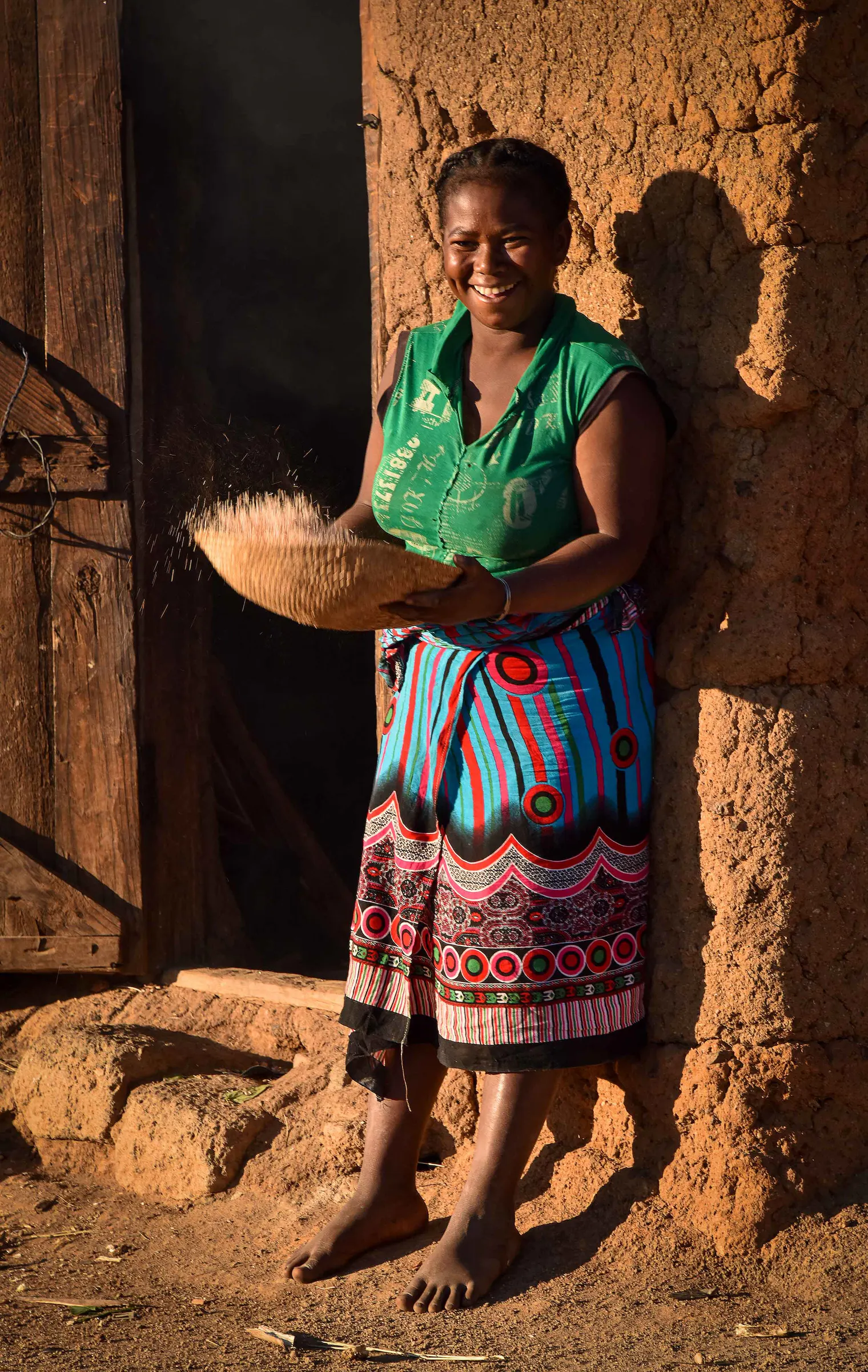 Une femme souriante vanne le riz avec un sahafa sur le seuil de sa maison